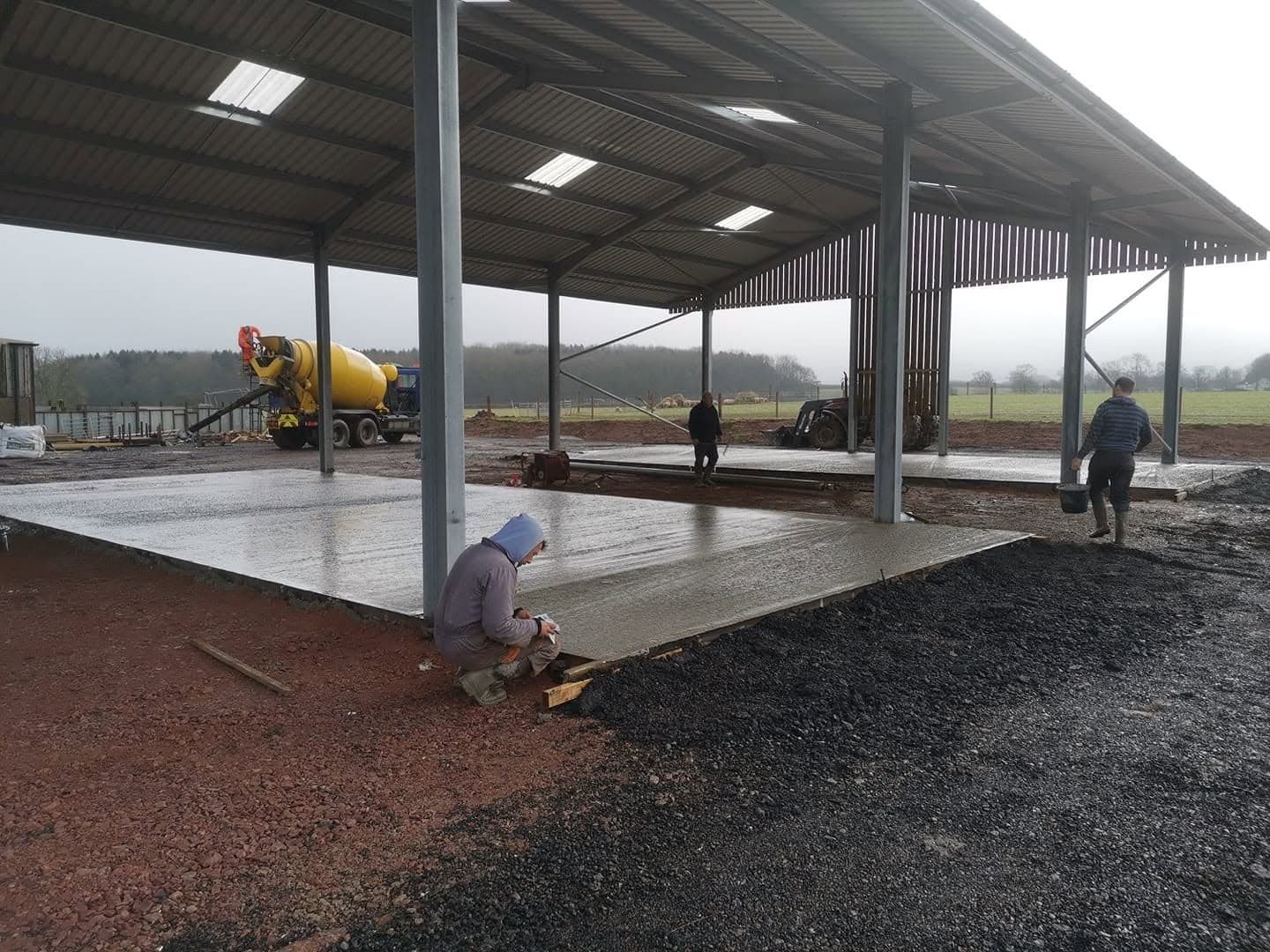 A concrete floor slab being laid inside a new steel-framed barn, with people working on the slab and a cement mixer lorry at the far end