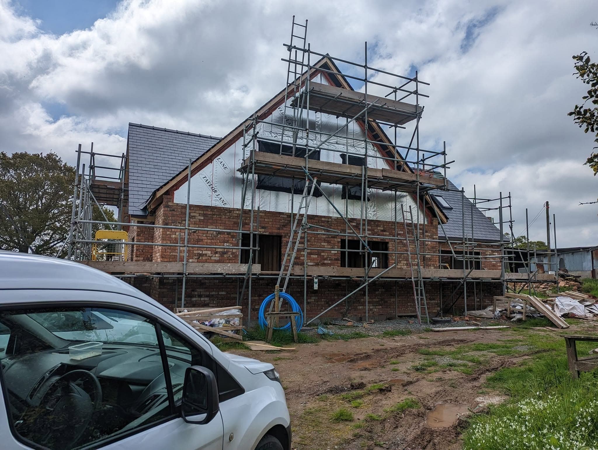 The gable end of a new brick house under scaffold, with a slate roof, roof windows and a white van parked in front
