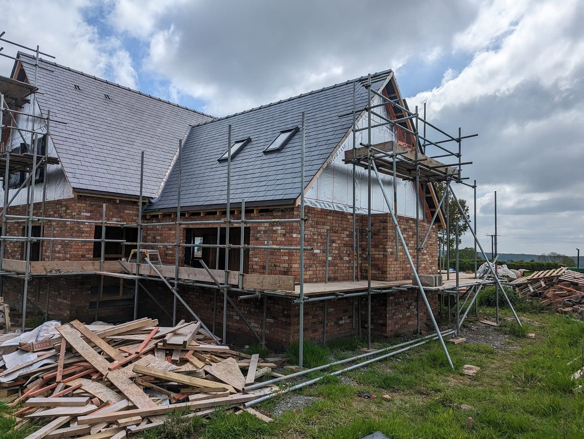 A new brick house with a slate roof and roof windows, with scaffolding around it and timber offcuts on the grass