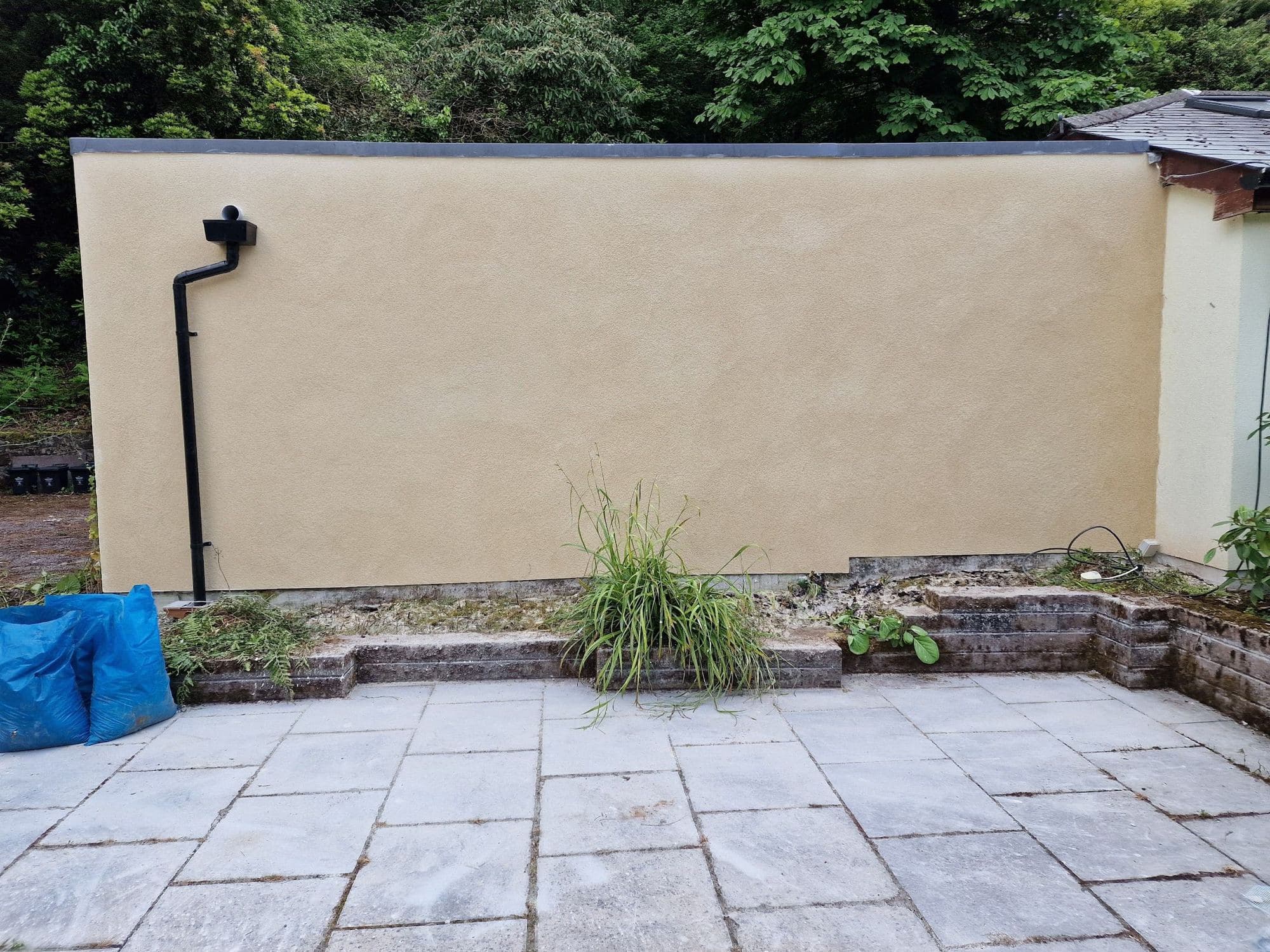 A smooth cream rendered wall with a black downpipe above a paved patio