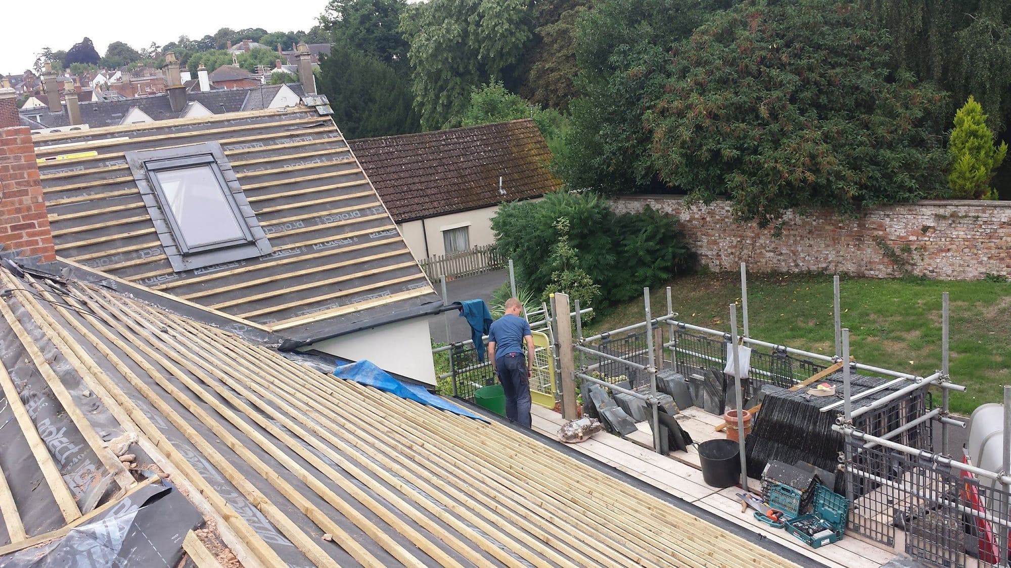 A roof stripped to battens with a roof window already set in, seen from the scaffold above a village