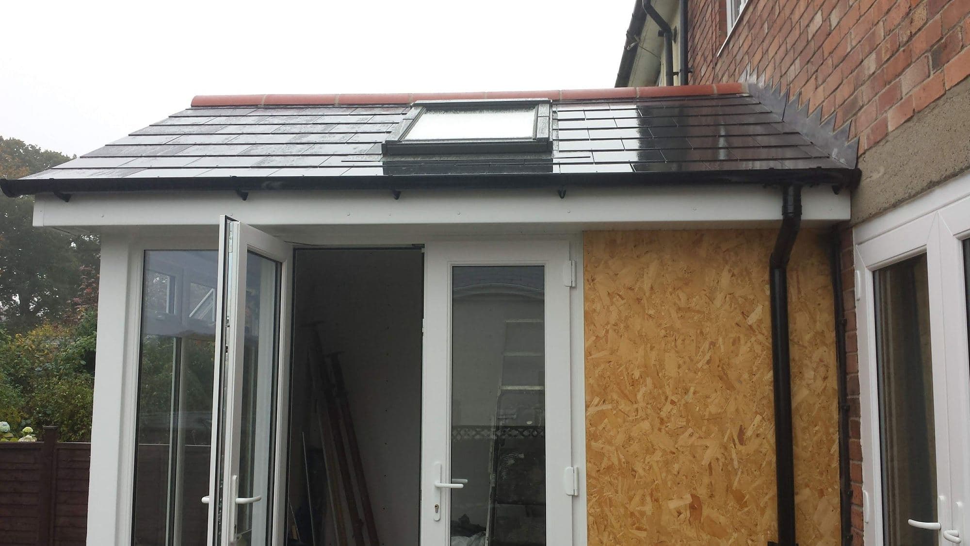 A small slate roof with a roof window above white double doors and a board-covered opening, against a brick house