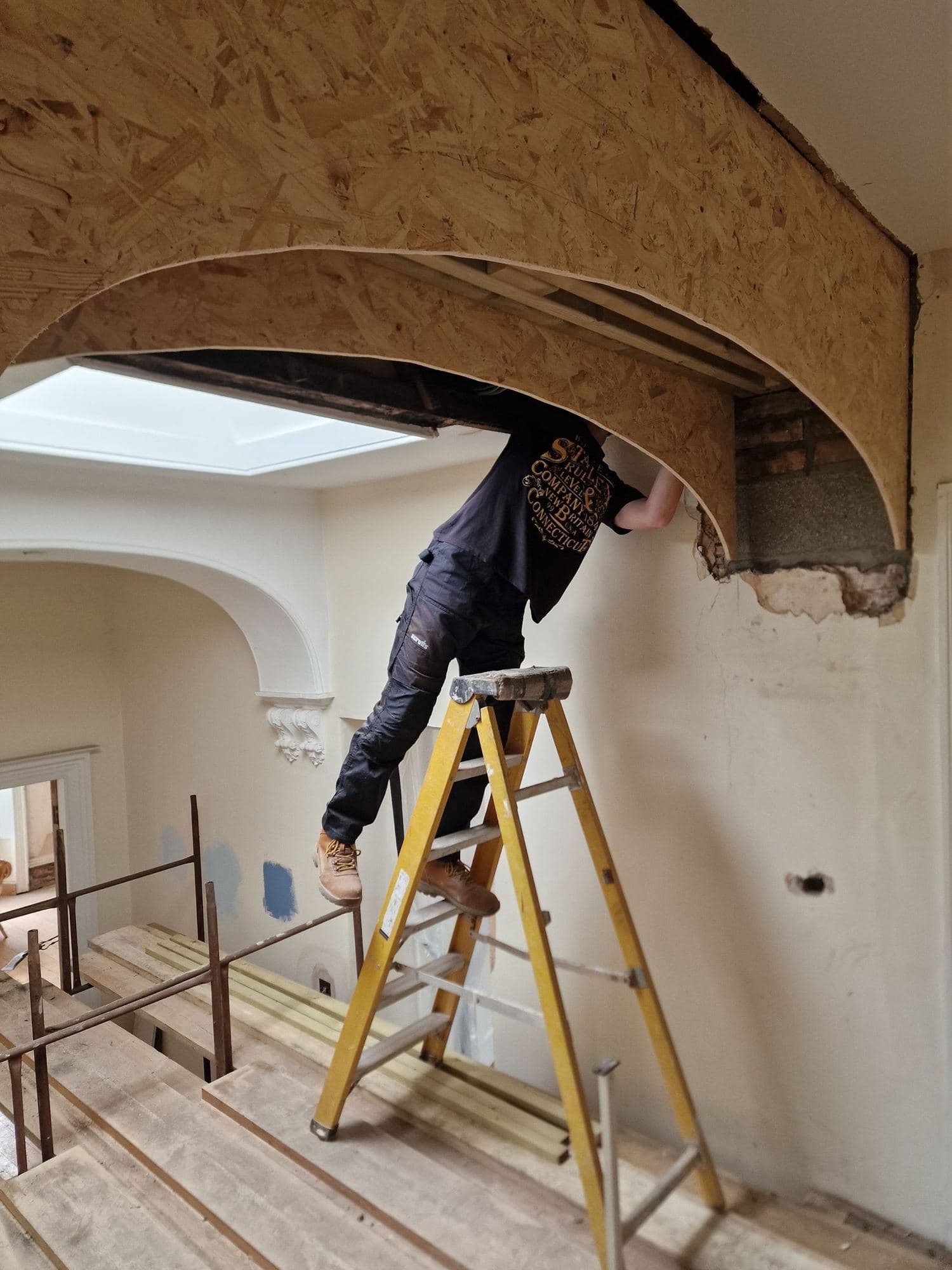 A man on a stepladder fitting a curved timber former for an arched opening in a hallway below a skylight