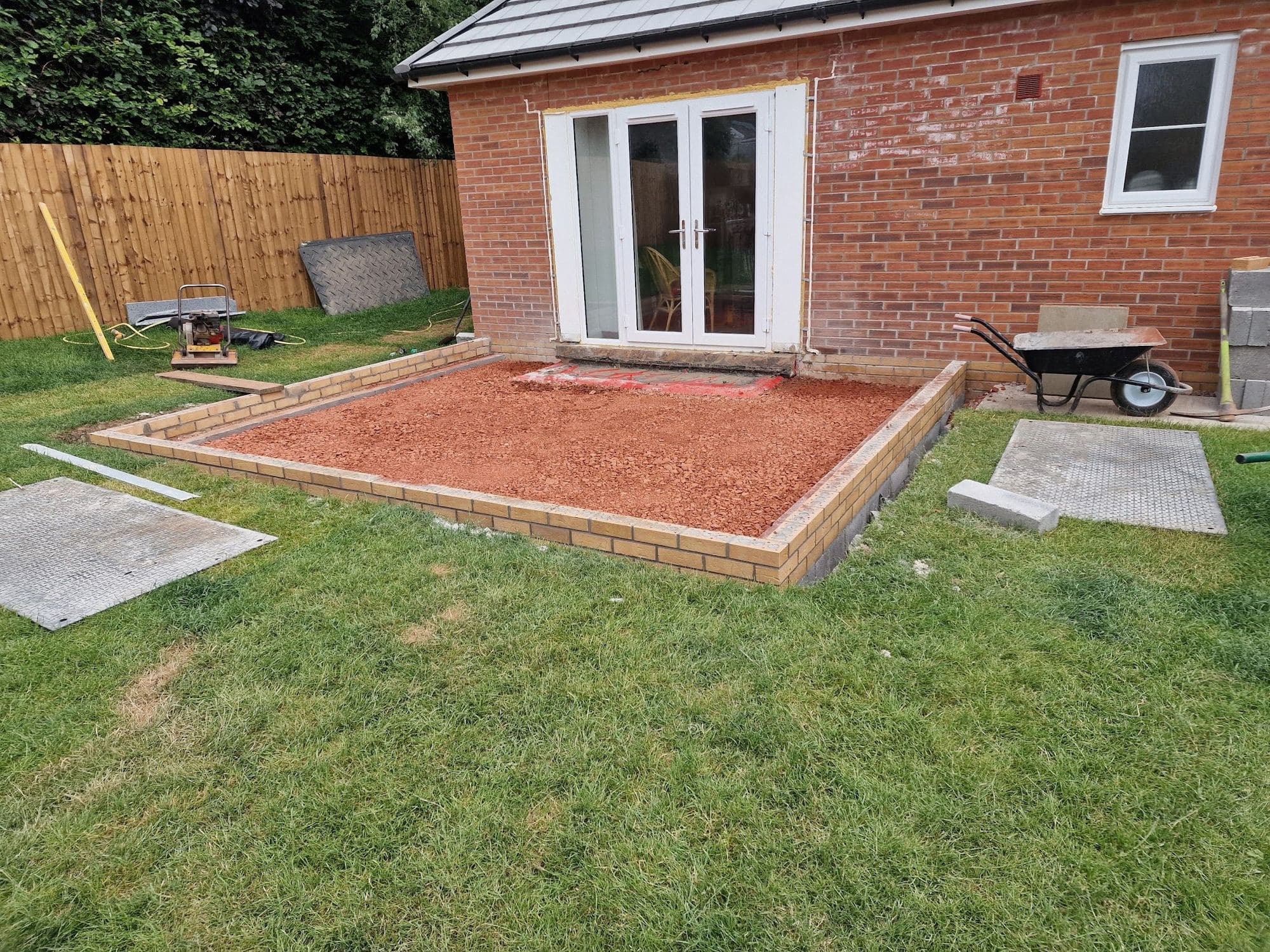 A brick and gravel base laid out for a new extension against the back of a red brick house with patio doors