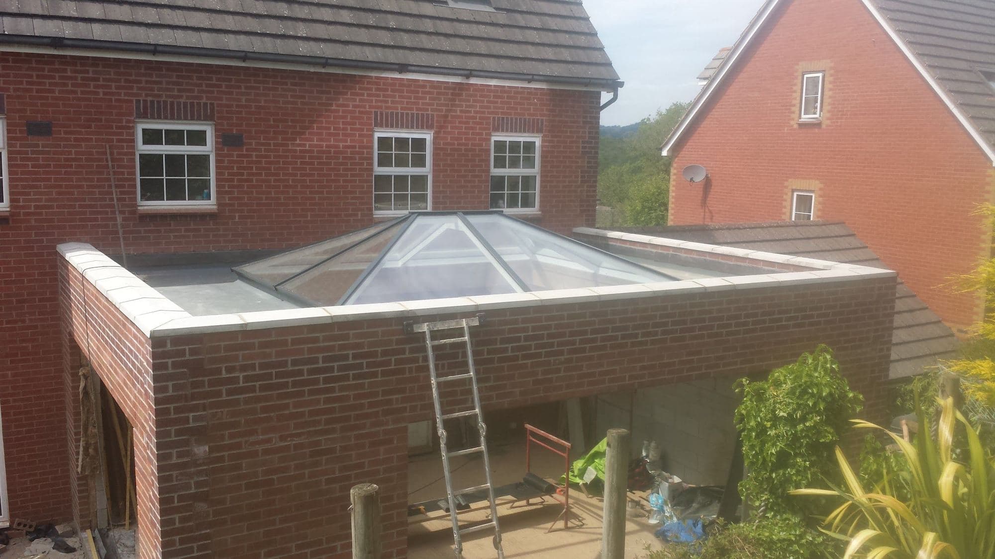 A brick extension with a glass roof lantern and a ladder against it, beside a red brick house