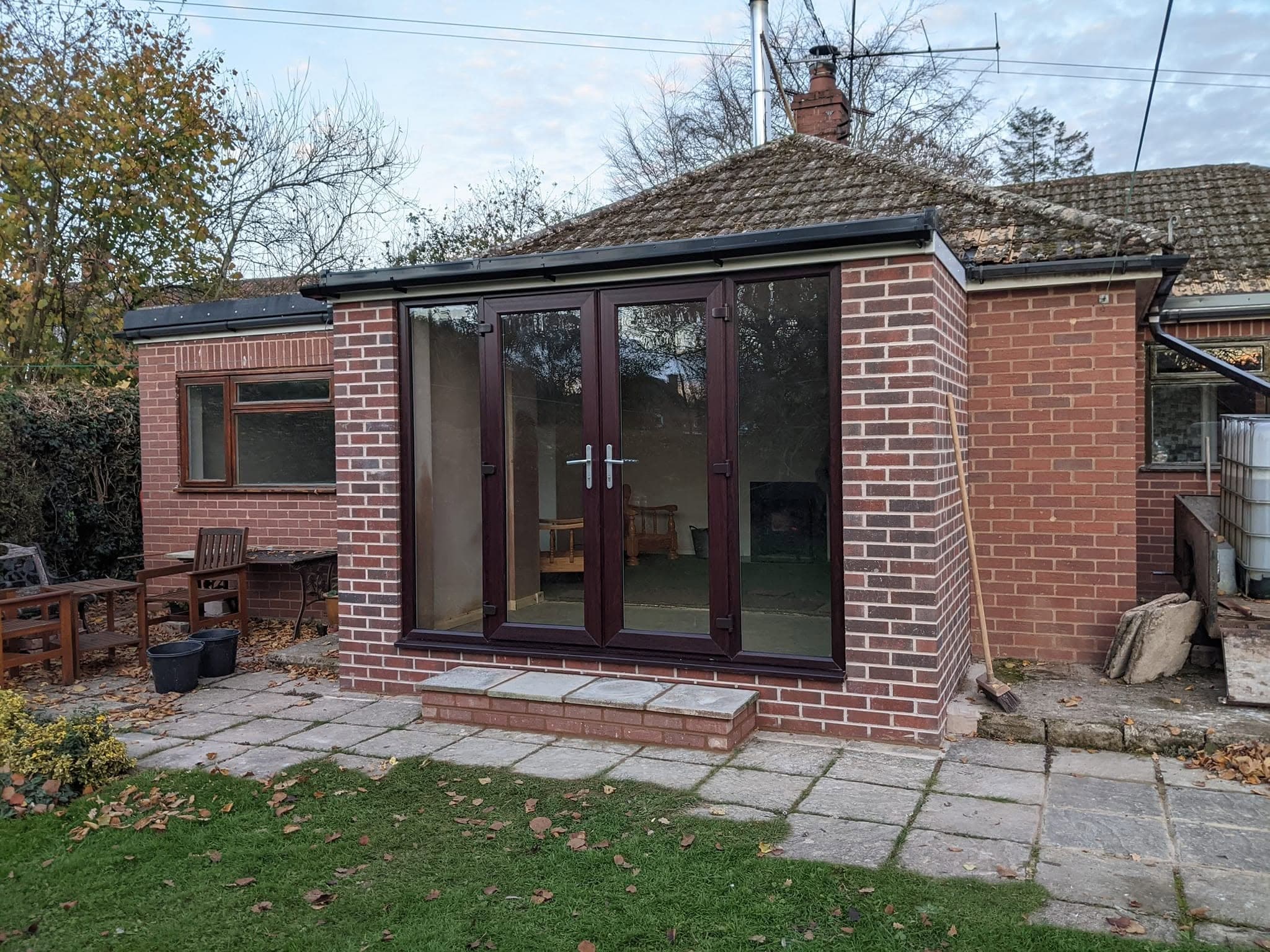 A single-storey brick extension with dark-framed patio doors and a tiled roof, on the back of a bungalow, with a paved patio and lawn in front