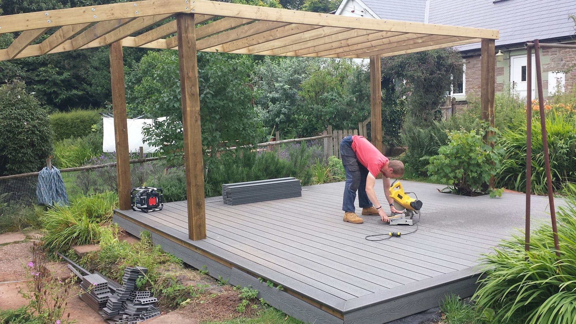 A man cutting decking boards with a chop saw beside a half-built timber pergola on a new deck in a planted garden