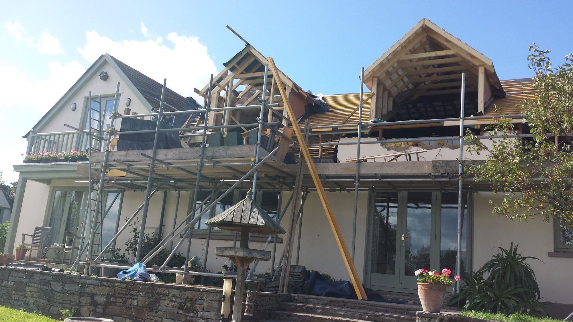 Two timber-framed dormers being built into the roof of a white house, with scaffolding across the front under a blue sky