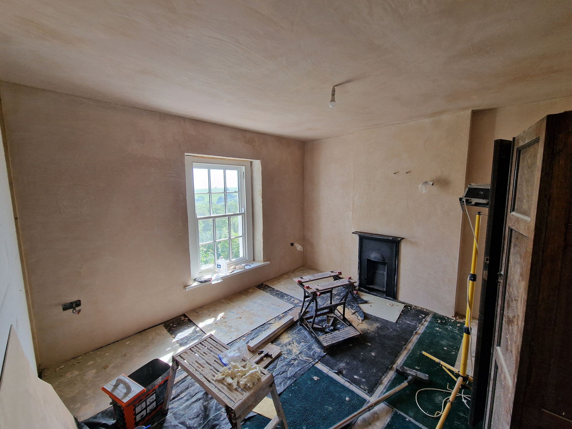 A bedroom with smooth new lime plaster walls and ceiling, a sash window looking onto trees and a cast iron fireplace