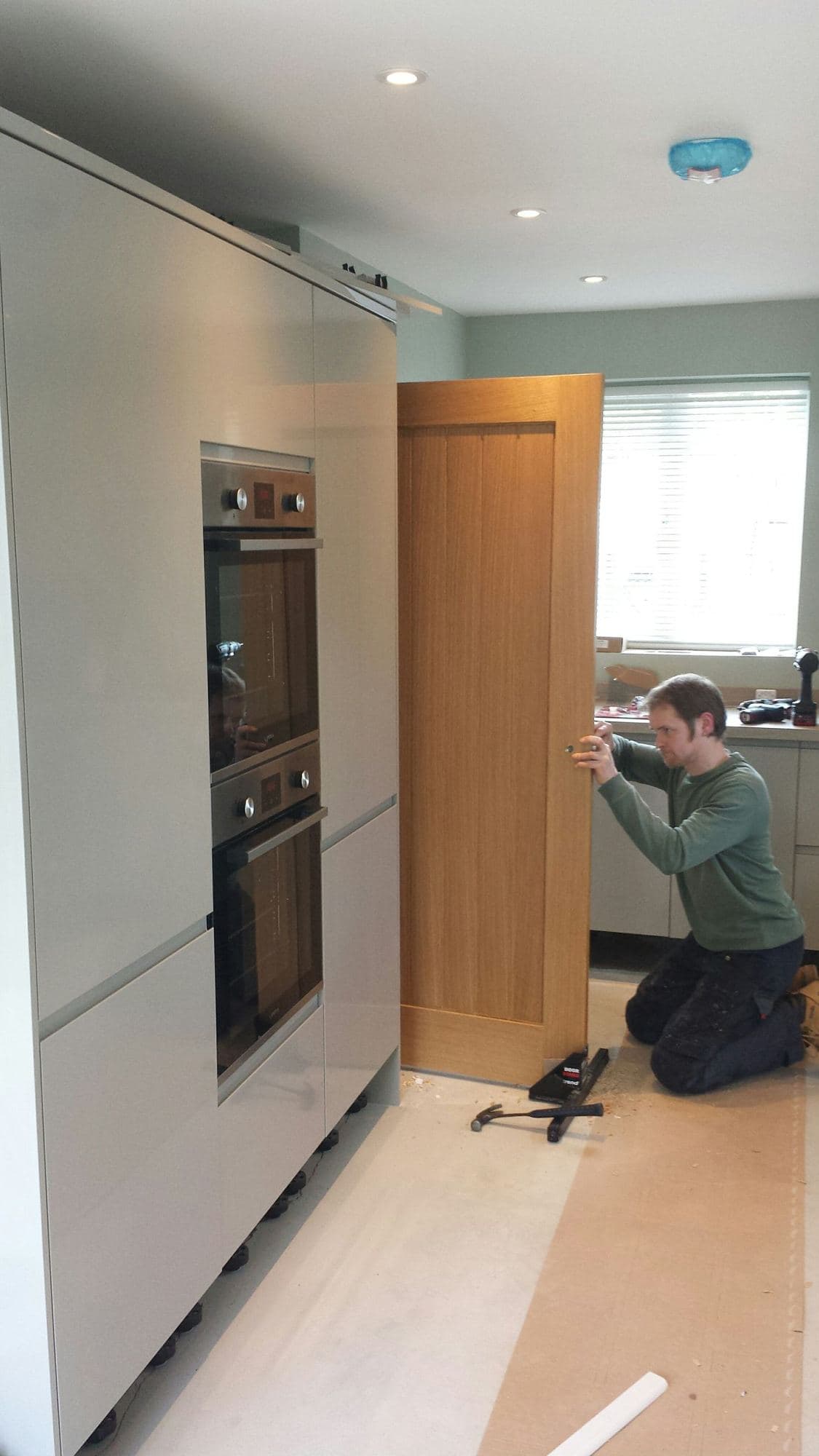 A man kneeling to fit an oak door beside tall kitchen units with a double oven