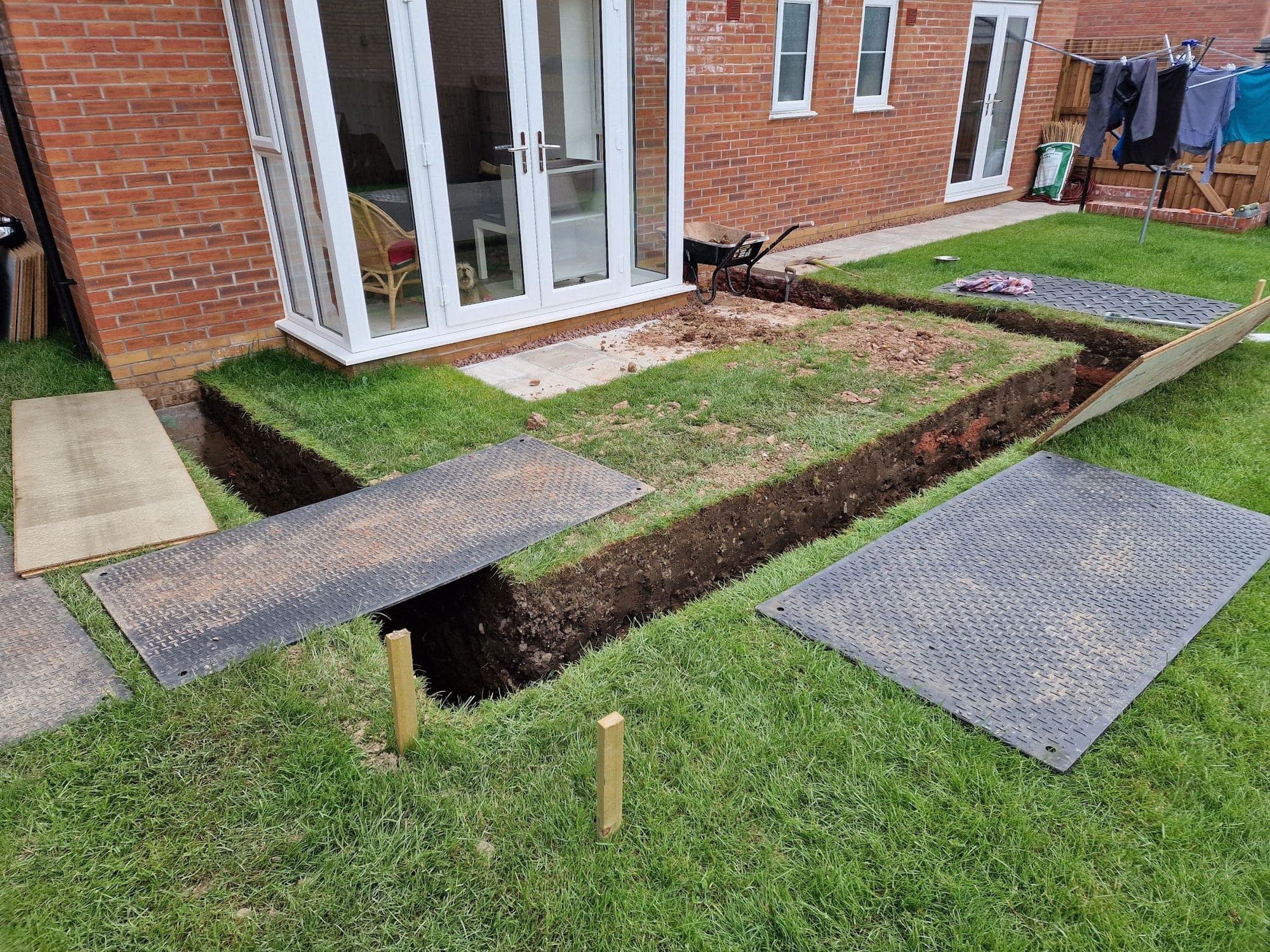 A narrow foundation trench dug across a lawn beside the back of a brick house, with ground mats and marker pegs