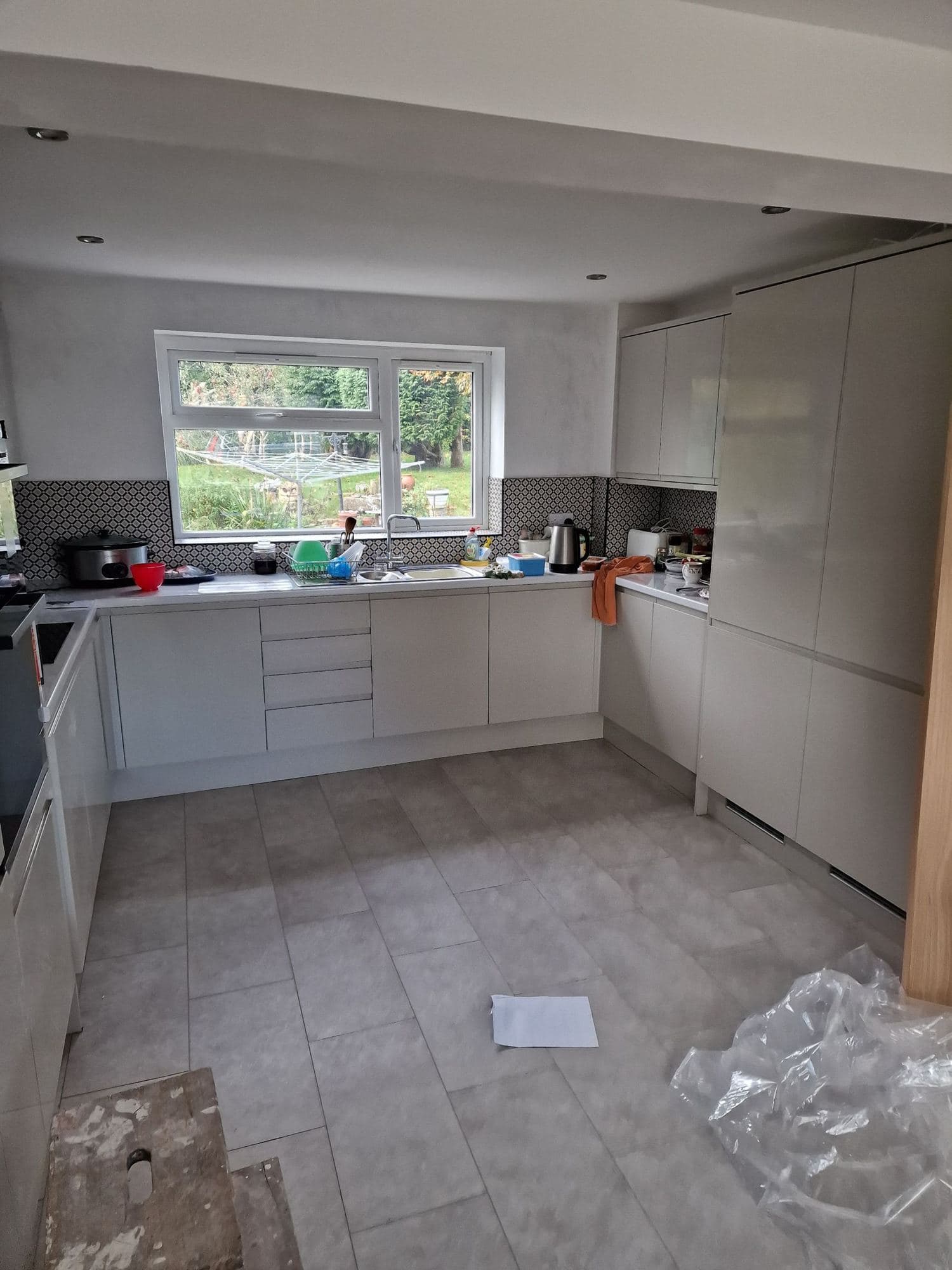A kitchen with pale handleless units, a window over the sink and a new tiled floor with off-cuts on it