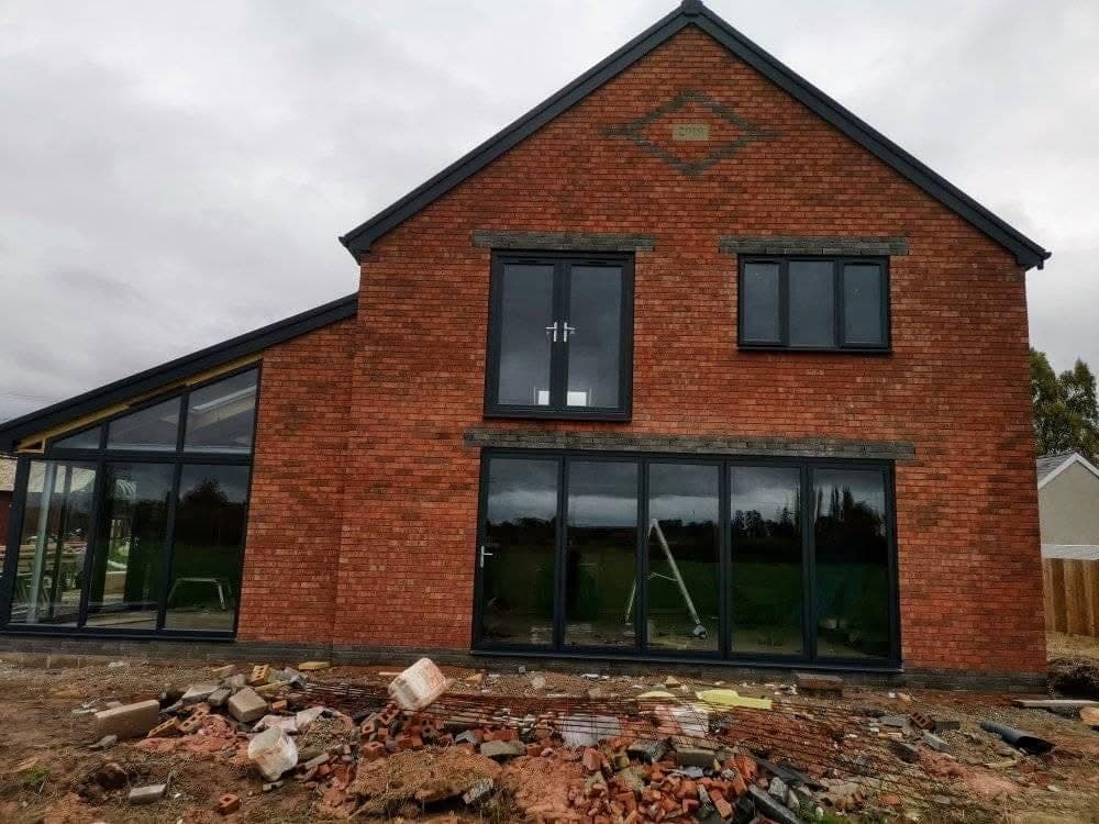 A new red-brick house with a gable end, dark-framed windows and bifold doors across the ground floor, and rubble on the ground in front