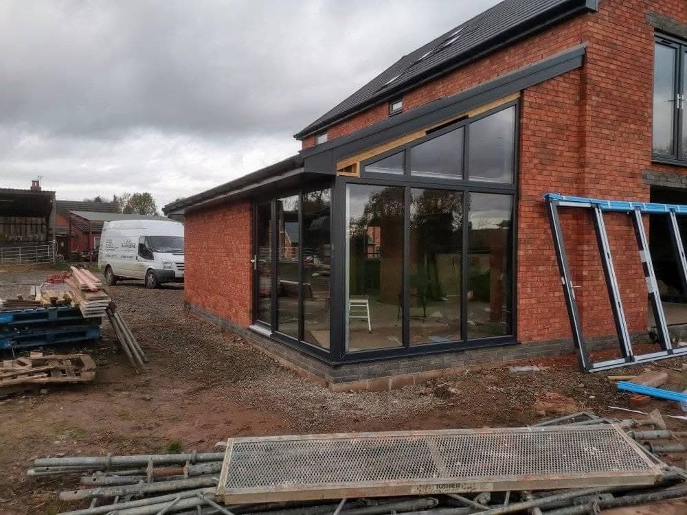 The corner of a new brick house with a glazed lean-to of dark-framed glass, and building materials stacked in front