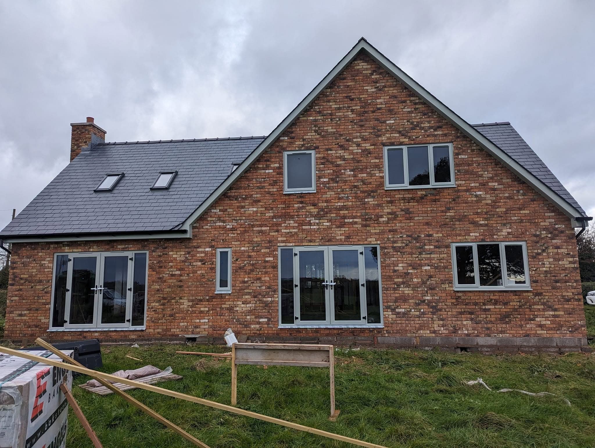 A new brick house with a slate roof, roof windows and grey-framed patio doors and windows, standing in a field