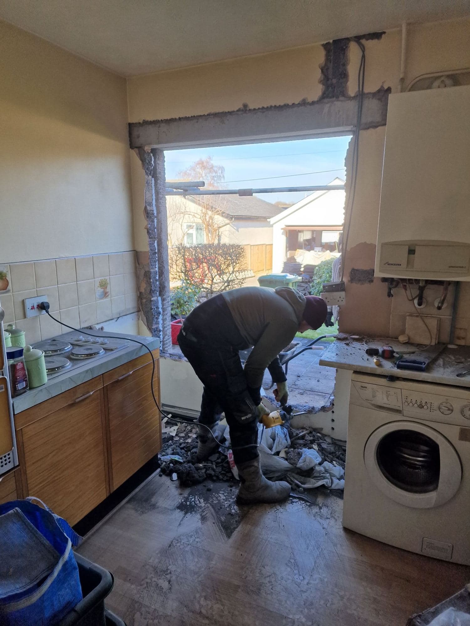 A man clearing rubble in an old kitchen beside a large new opening cut in the outside wall
