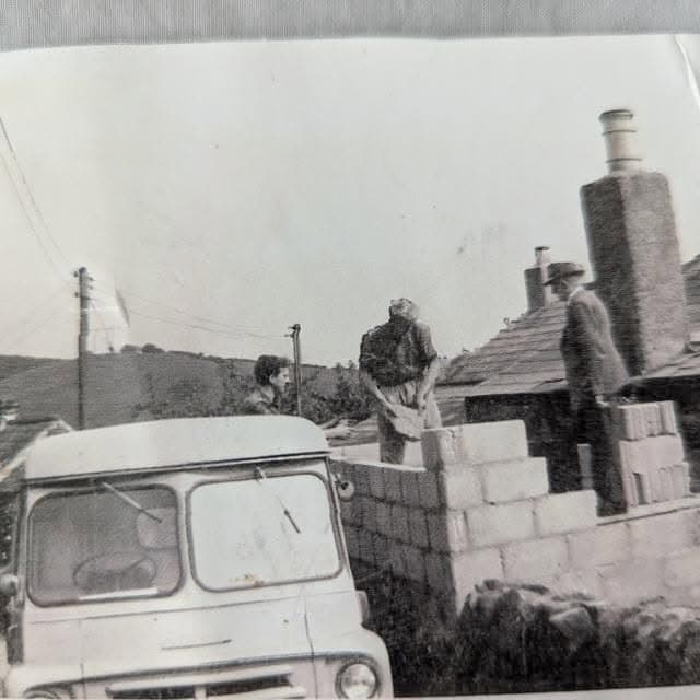 An old black and white photo of three men building a blockwork wall beside a cottage roof, with a van in the foreground