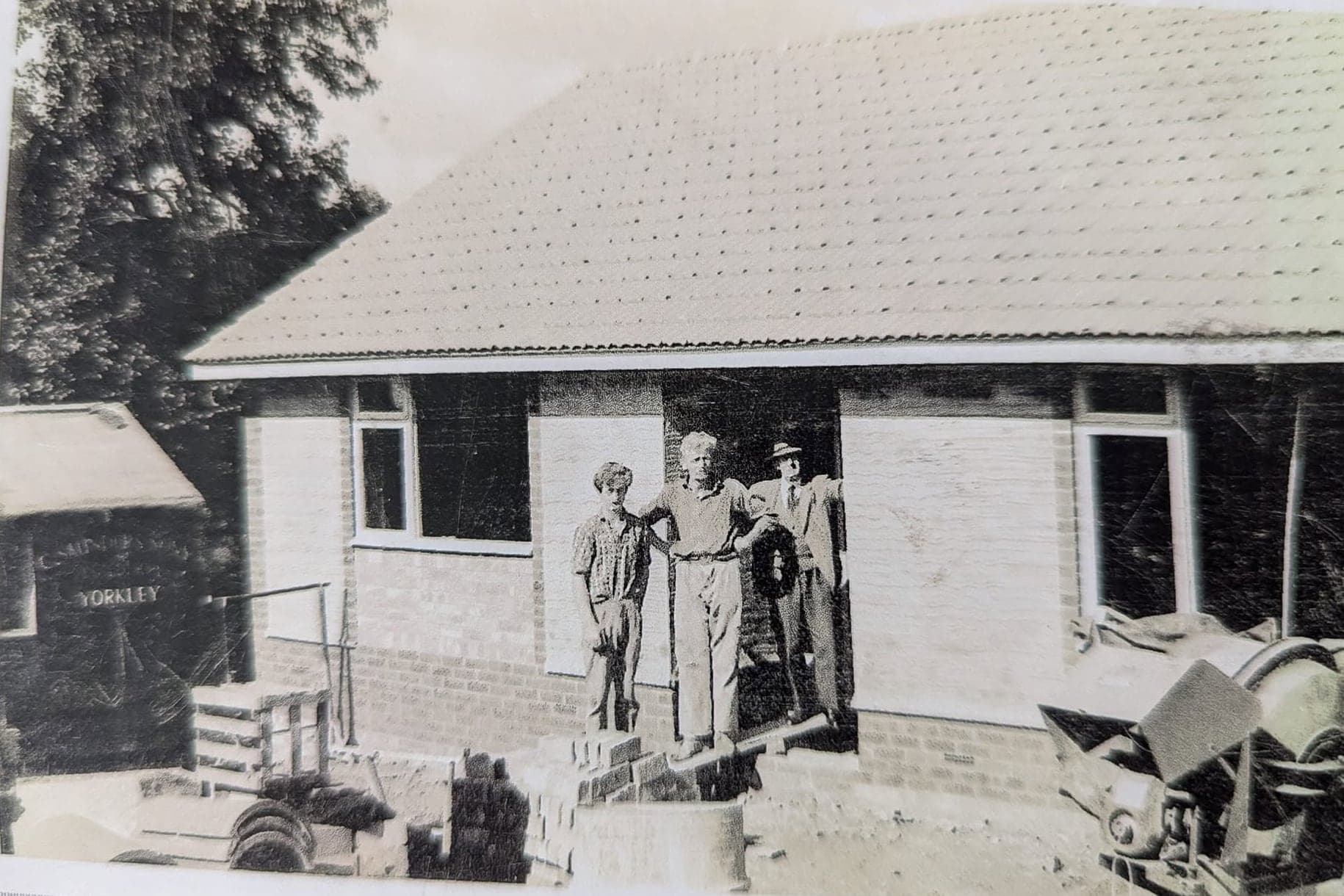 An old black and white photo of three men standing outside a half-built brick bungalow, with a cement mixer on the right