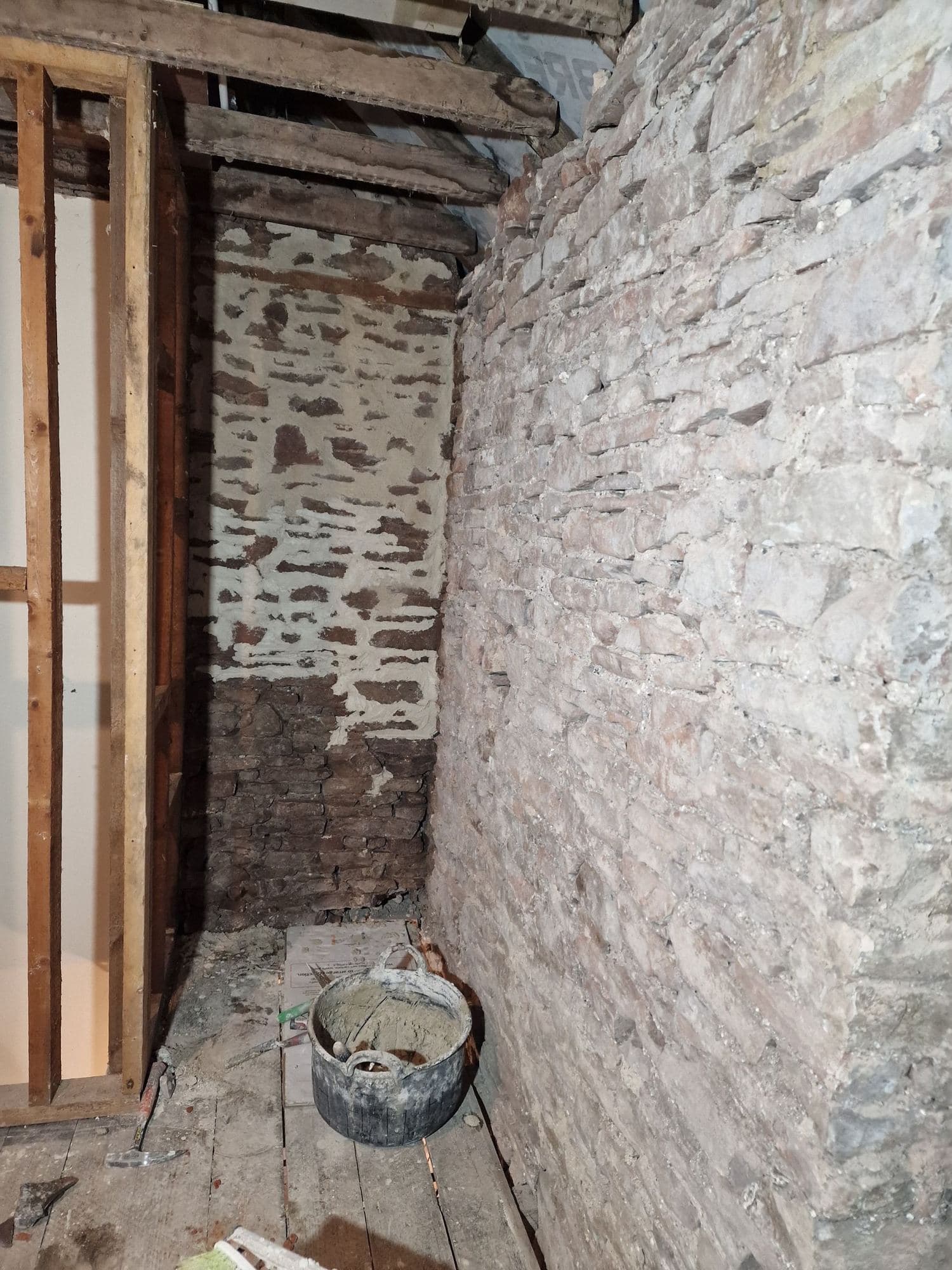 An old timber stud partition and a rough stone wall stripped back in a narrow room, with a bucket on the floor