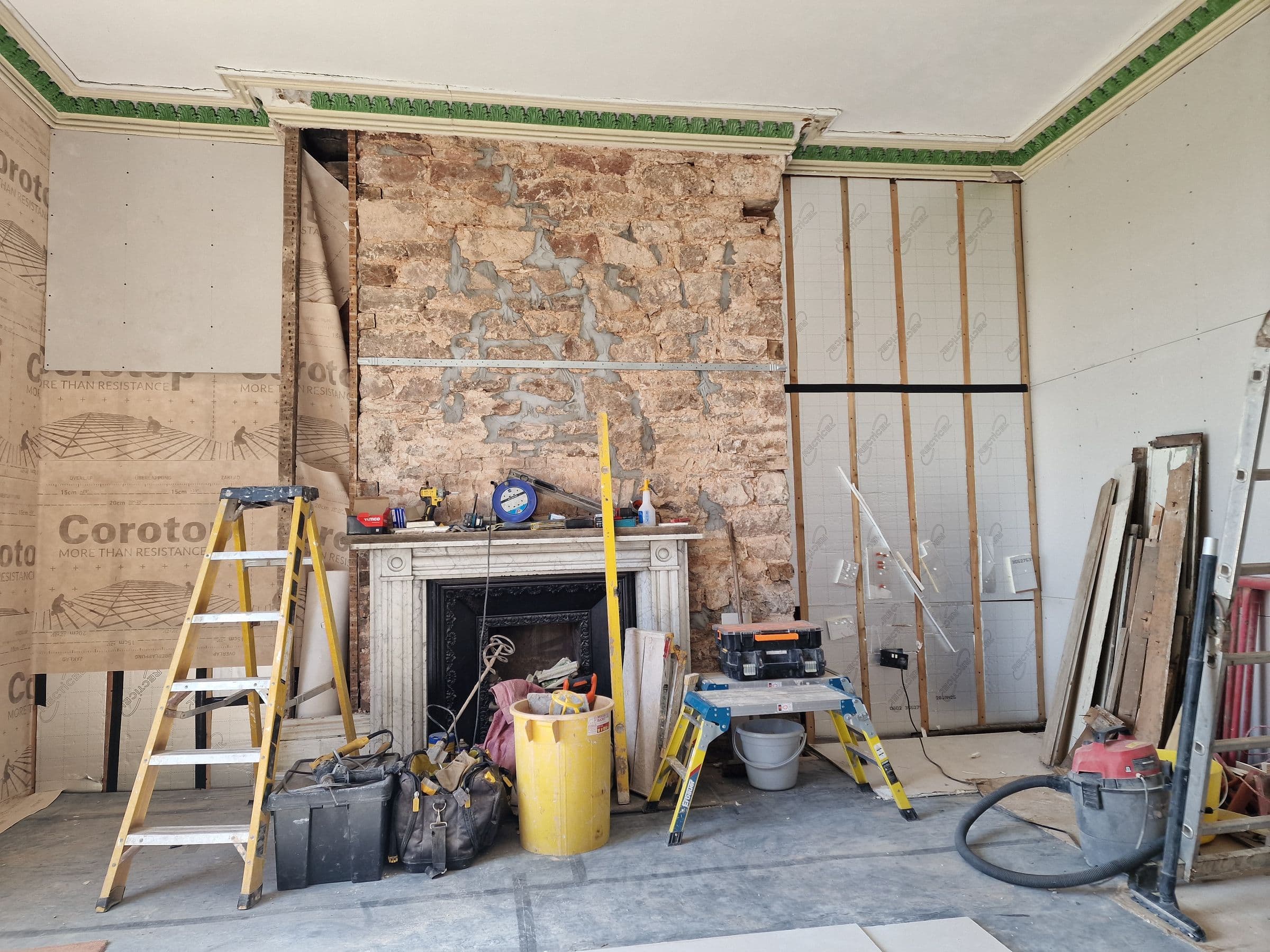 A high-ceilinged period room with the chimney breast stripped back to stone, original cornice kept, and walls being boarded