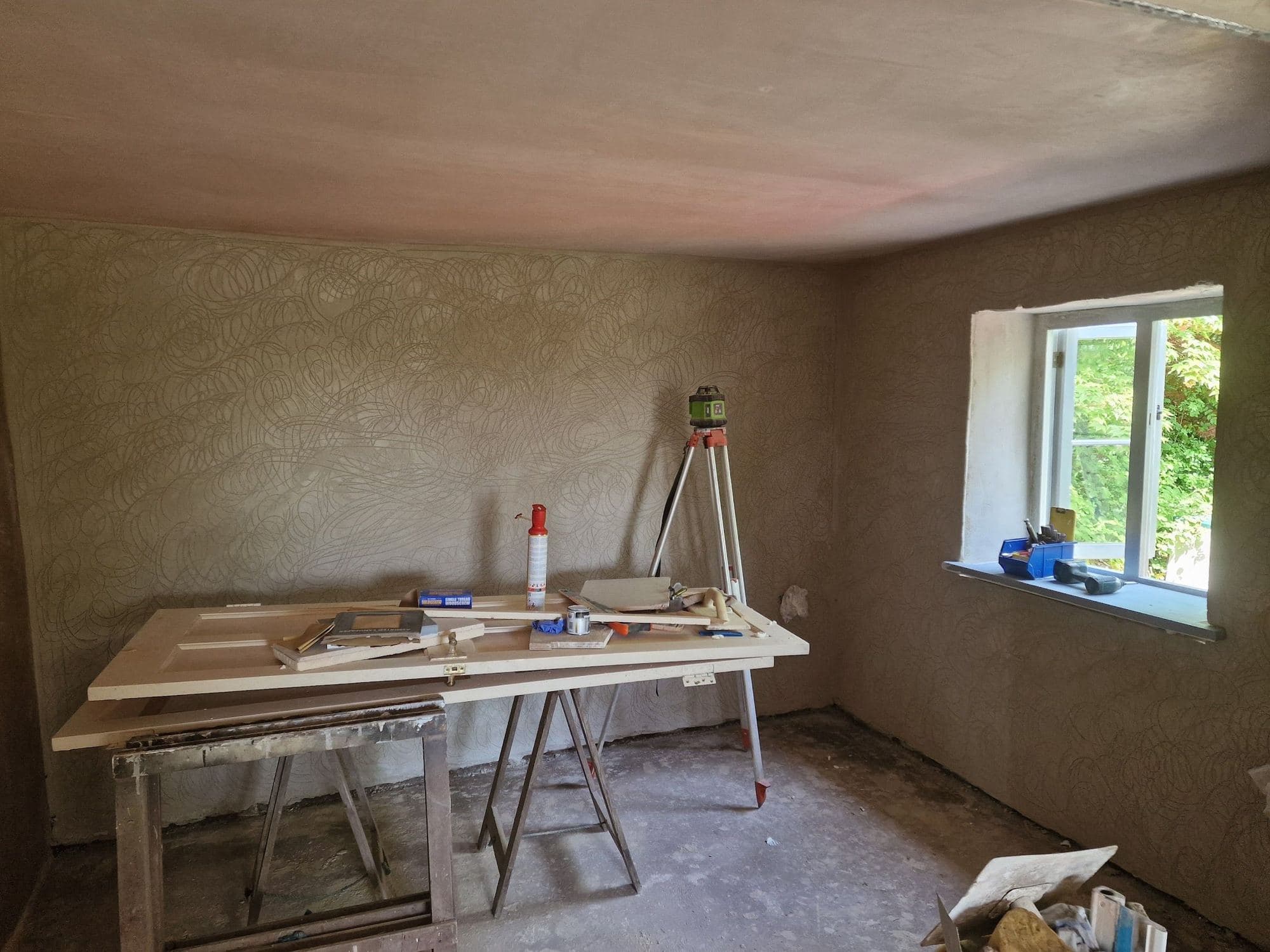 A newly plastered room with a window, a trestle table with timber on it and a laser level on a tripod