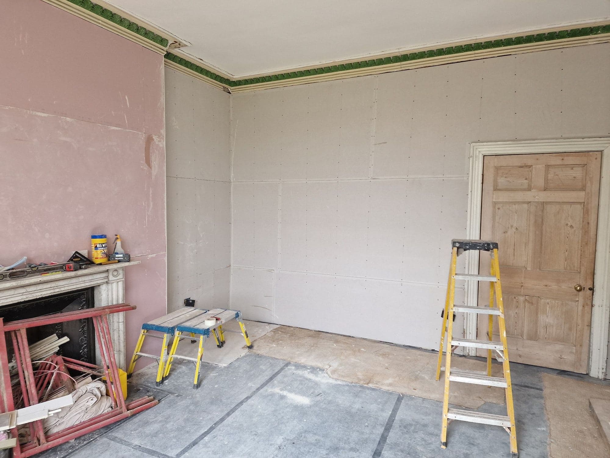 A room with plasterboard fixed to the walls, a green cornice kept at the top and a panelled door