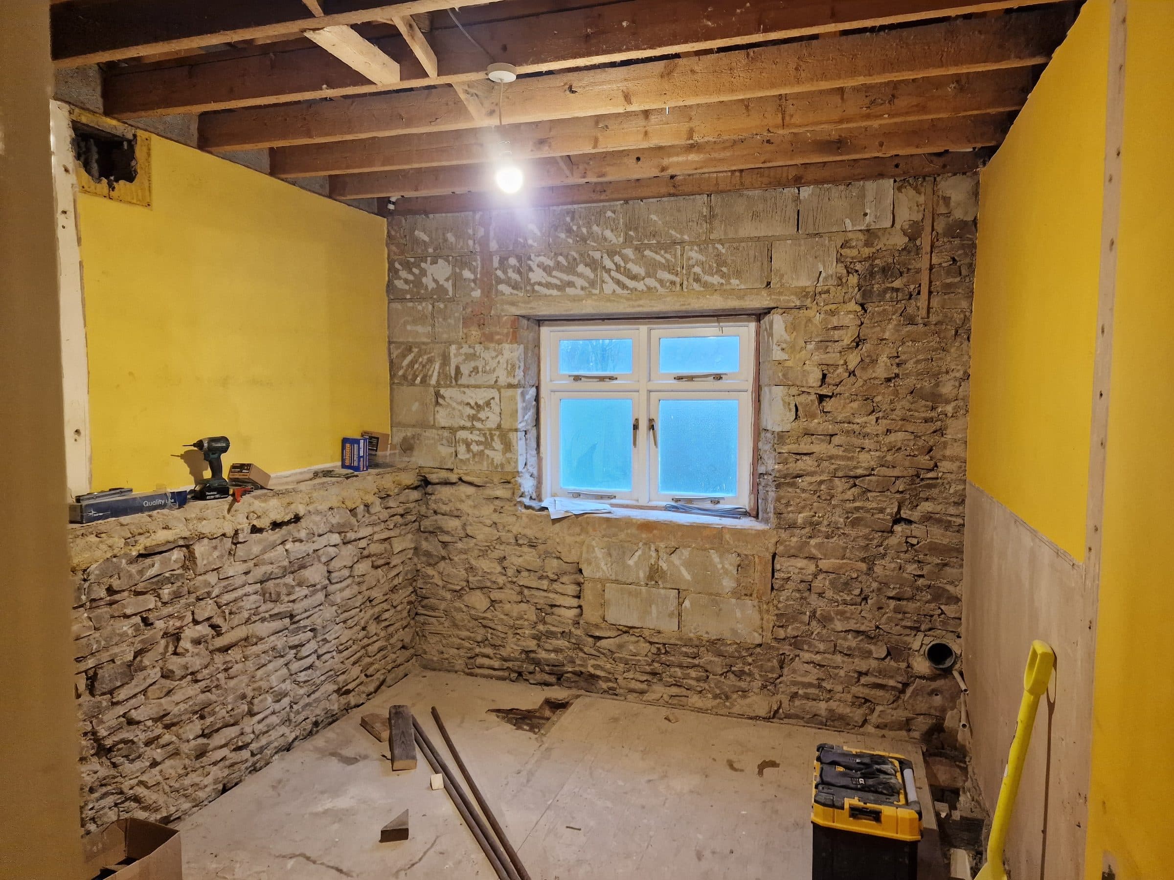 A cottage room stripped back to its rubble and dressed stone walls, with the joists exposed and a white casement window