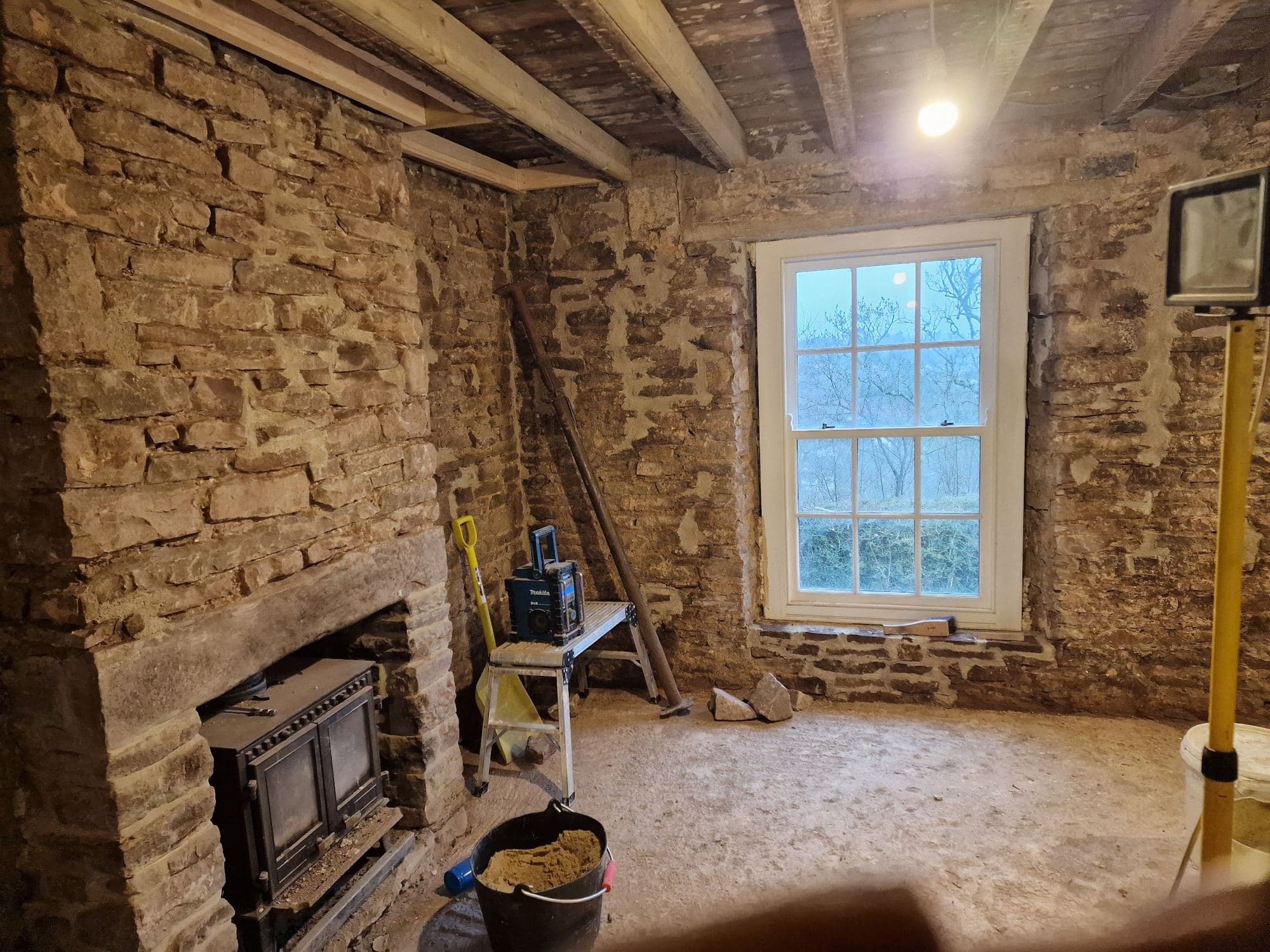 A stone-walled room with a sash window, a stone chimney breast and a wood stove, and tools on the floor