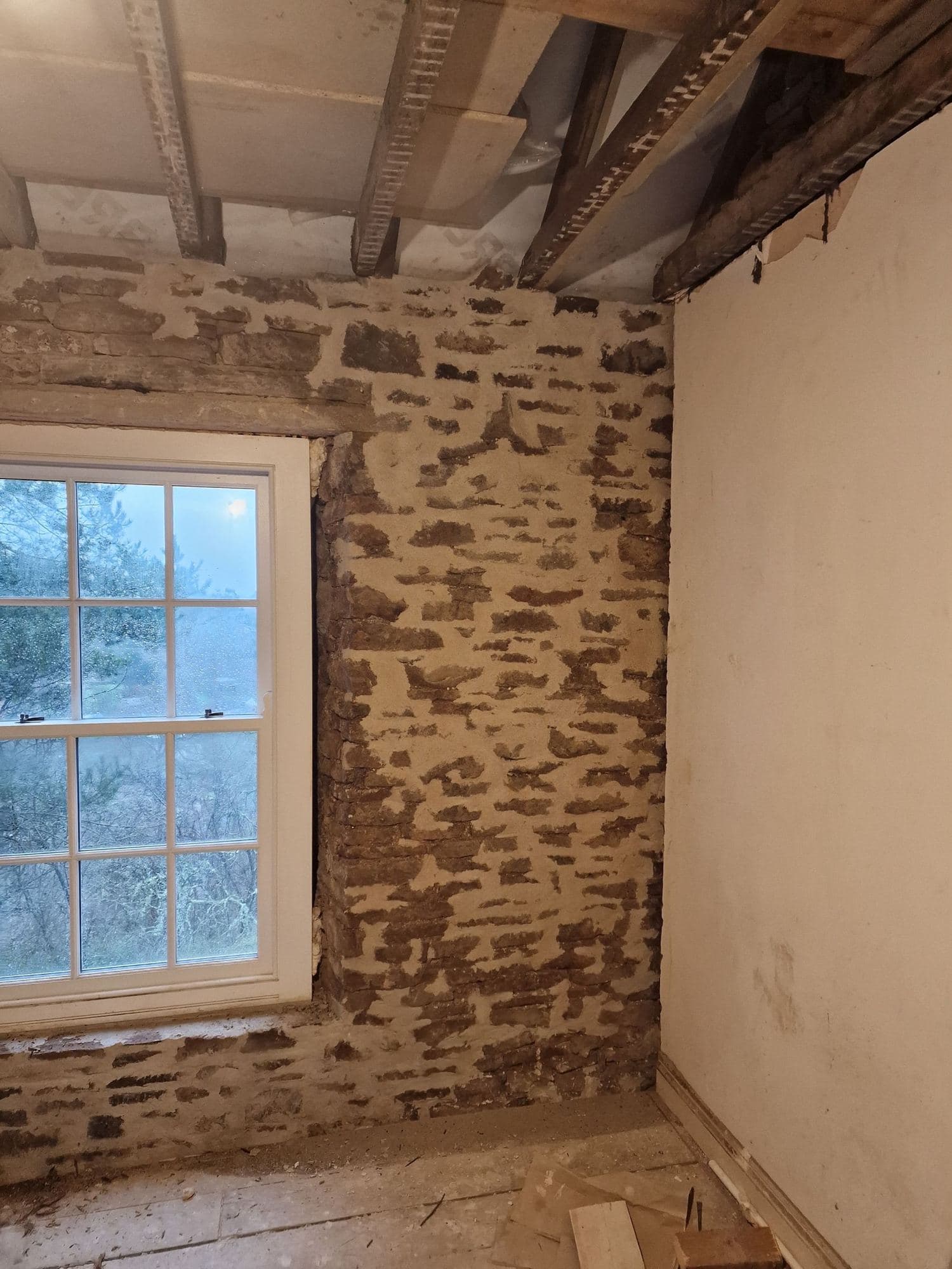A room stripped back to rubble stone with a white sash window and exposed joists above