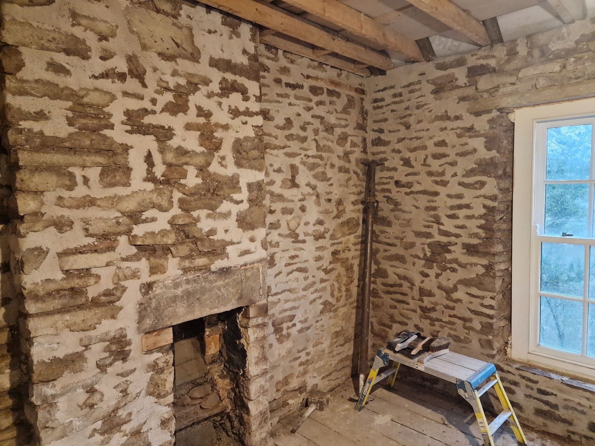 Rubble stone walls patched with pale mortar either side of a fireplace opening, with a sash window to one side