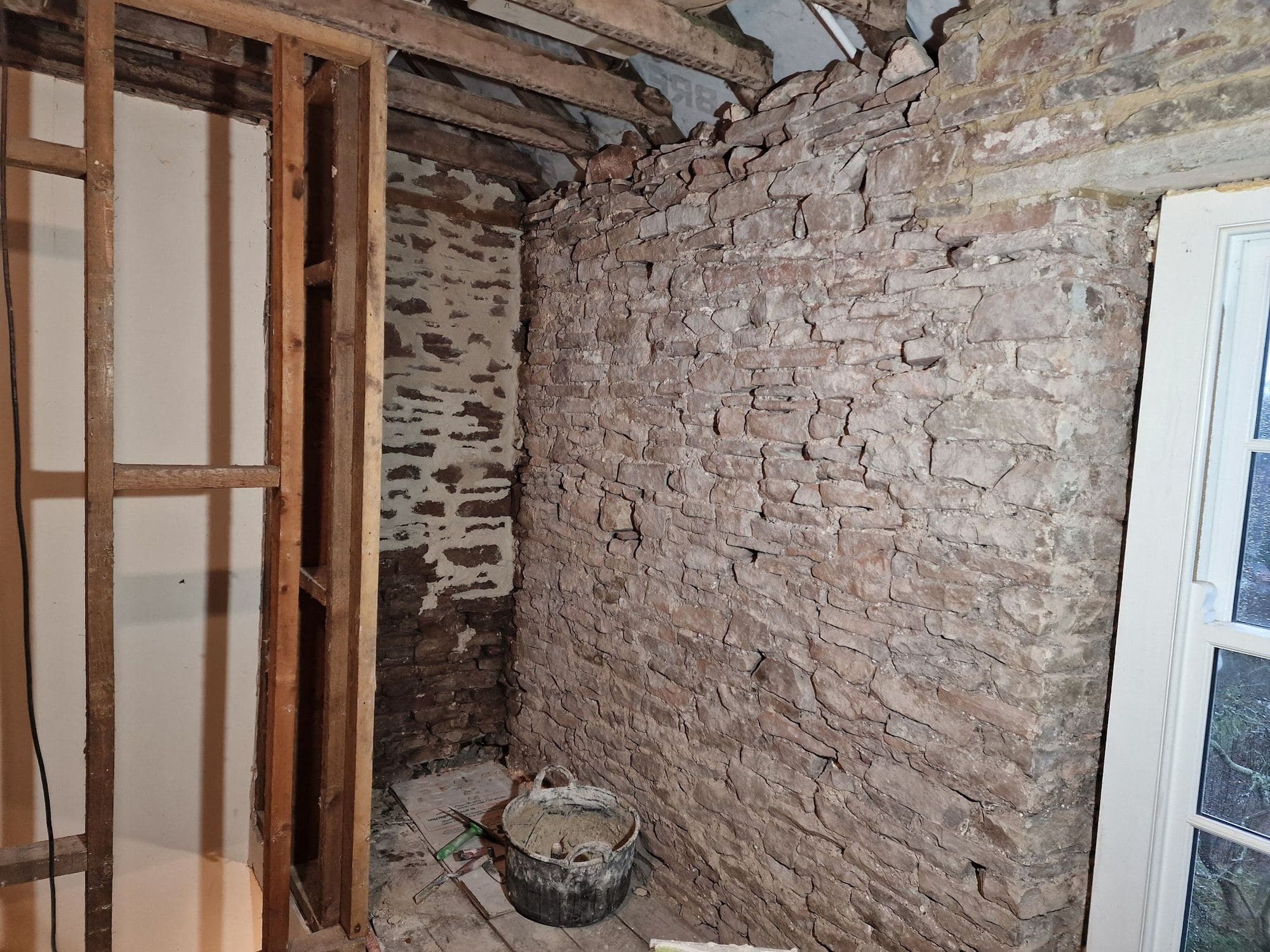 New timber studwork standing beside a rubble stone wall in an old room, with joists showing above